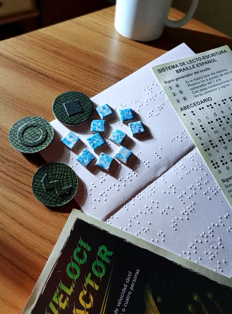 Escena sobre una mesa de madera con componentes del juego Velocitactor. Se ven tres fichas redondas en relieve con figuras geométricas, varias fichas cuadradas azules de puntuación, el reglamento ilustrado, el reglamento en braille y una hoja impresa con el sistema de lectoescritura braille en español. Al fondo aparece una taza blanca de café como parte decorativa.