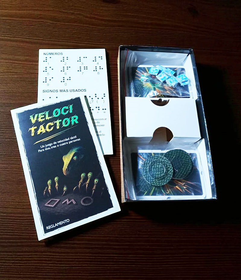 Caja abierta del juego Velocitactor con los componentes organizados en compartimentos. Se distinguen los mazos de cartas en bolsas plásticas, las fichas redondas y las fichas azules de puntuación. A la izquierda, sobre la mesa, está el reglamento ilustrado con la portada visible y detrás una hoja en braille con números y signos más usados.